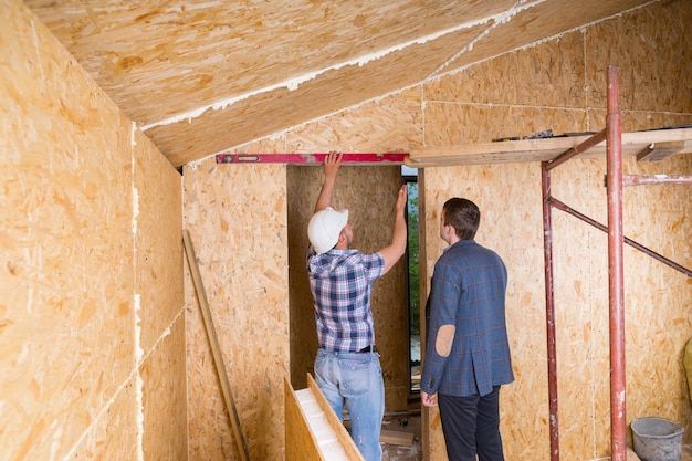 Baumeister und Architekt bei der Inspektion der Konstruktion des Türrahmens mit einer Ebene im Inneren des unfertigen Hauses mit freiliegenden Sperrholzplatten