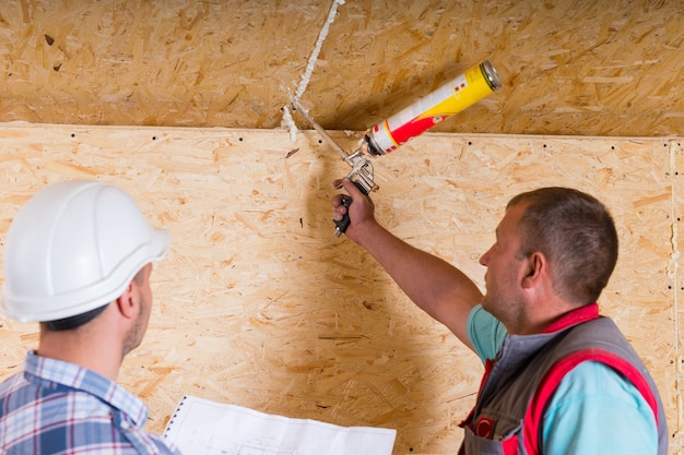 Baustellenvorarbeiter mit weißem Schutzhelm, der Pläne hält und Arbeiter beim Abdichten auf unfertige Holzdecke beobachtet