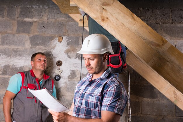 Baustellenvorarbeiter mit weißem Schutzhelm und Bauplänen vor dem Team von Arbeitern, die eine Holztreppe im unvollendeten Keller des neuen Hauses bauen