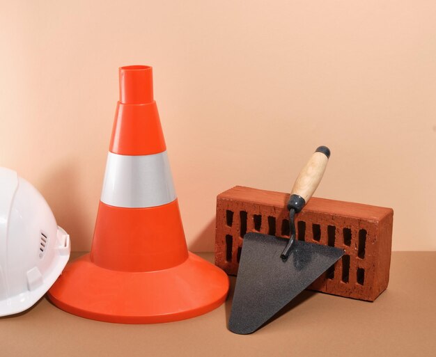 Bautechnische Werkzeuge auf einem beigenen Hintergrund Baukegel und weißer Kopfschutzhelm orangefarbener Ziegelstein und Trowel