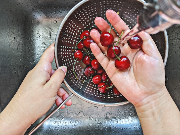 La baya de cerezas frescas maduras en un tamiz de acero se lava a mano con agua del grifo en el fregadero, vista superior