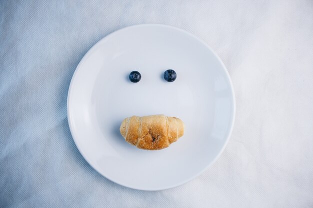 Bayas frescas de arándano y croissant en forma de una cara con una sonrisa en un plato blanco.