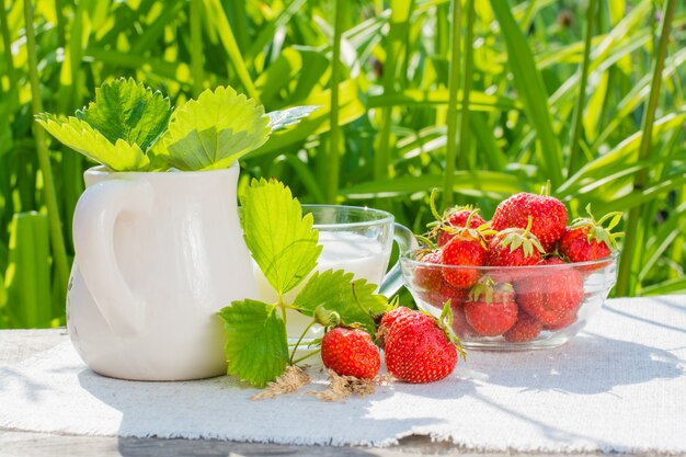 Bayas y hojas de fresa, un tazón de fresas, una jarra y una taza de leche en una servilleta sobre una mesa de madera