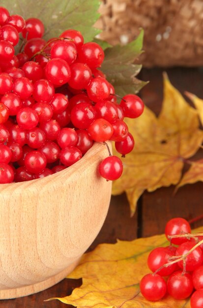 Bayas rojas de viburnum en tazón de madera con hojas amarillas en la mesa sobre fondo marrón