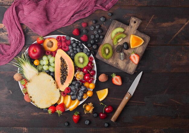 Bayas de verano orgánicas frescas y frutas exóticas en plato blanco sobre fondo de madera oscura con tabla de cortar y cuchillo. Piña, papaya, uva, nectarina, naranja, albaricoque, kiwi, pera. Vista superior