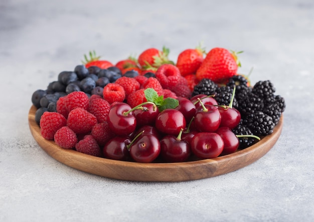 Las bayas de verano orgánicas frescas se mezclan en una bandeja redonda de madera sobre un fondo claro de mesa de cocina Frambuesas fresas arándanos moras y cerezas Vista superior