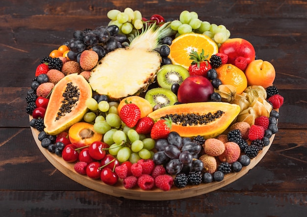 Bayas de verano orgánicos crudos frescos y frutas exóticas en bandeja grande redonda sobre fondo de cocina de madera oscura. Papaya, uva, nectarina, naranja, frambuesa, kiwi, fresa, lichis, cereza. Vista superior