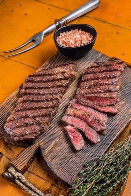 BBQ gegrilltes Denver Top Blade Beefsteak mit Thymian auf einem Holzbrett Orangefarbener Hintergrund Ansicht von oben