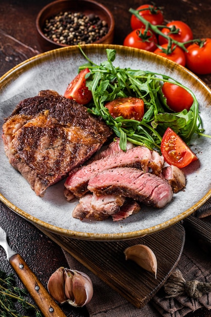 BBQ Gegrilltes Rib-Eye-Steak gebratenes Ribeye-Rindfleisch auf einem Teller mit grünem Salat Draufsicht auf dunklen Hintergrund