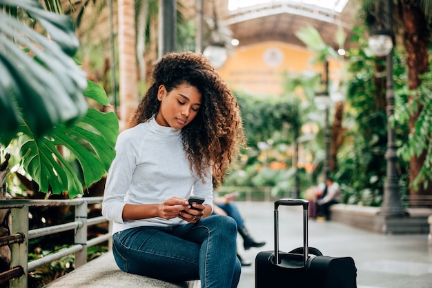 Beaituful weiblicher Tourist mit dem gelockten Haar unter Verwendung des Smartphone. Draußen sitzen mit Koffer.