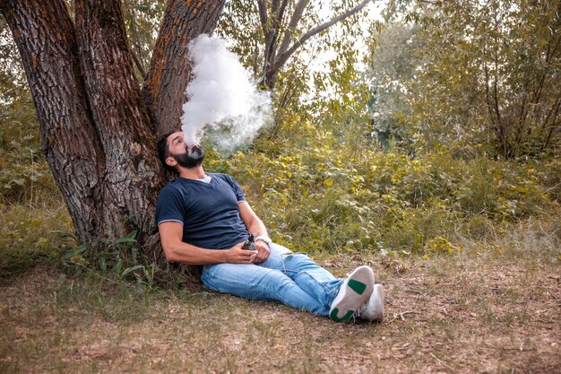 Beardedsmoker está descansando en el bosque y vapeando y desahogándose con un cigarrillo electrónico. Deshacerse de la adicción a la nicotina.