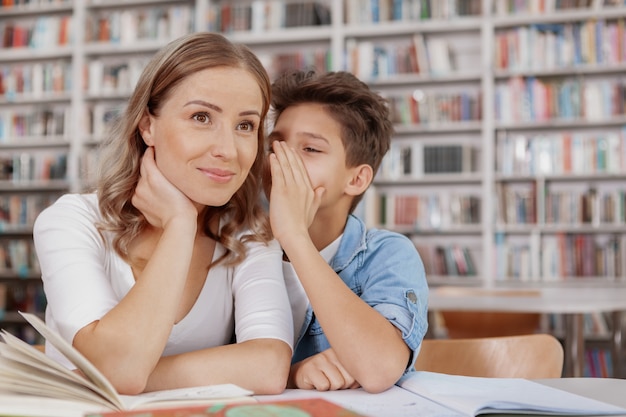 Beauitful mujer sonriendo cuando su pequeño hijo le susurra al oído en la biblioteca