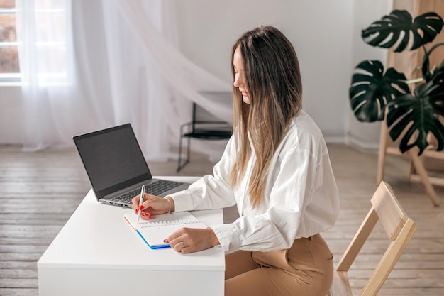 Beauitul jovem mulher trabalhando usando laptop concentrado e sorrindo e escreve em um caderno