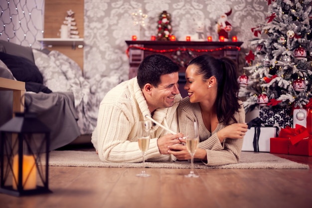 Beautifl joven marido y mujer que se establecen en el suelo riendo el día de Navidad. Pareja en sala de estar.