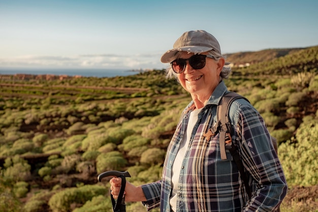 Beautiful senior Woman in Hut und Freizeitkleidung Wandern in der Natur genießen Freiheit und Urlaub Happy reife Frau Blick in die Kamera lächelnd Berg und Meer im Hintergrund