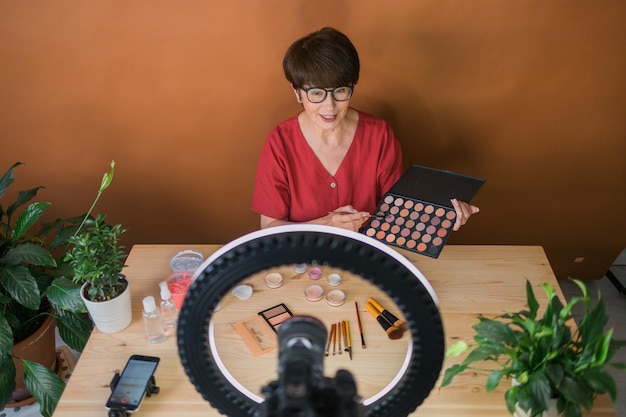 Beauty-Bloggerin oder Influencerin mittleren Alters mit Make-up-Produktaufnahmevideo im Zimmer zu Hause Mit Ringlampe und Smartphone