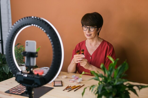 Beauty-Bloggerin oder Influencerin mittleren Alters mit Make-up-Produktaufnahmevideo im Zimmer zu Hause Mit Ringlampe und Smartphone