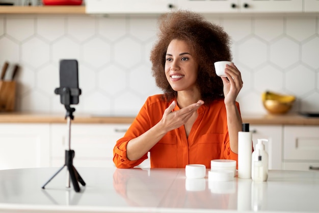 Beauty Blogging Mujer negra sonriente revisando productos para el cuidado de la piel frente a la cámara