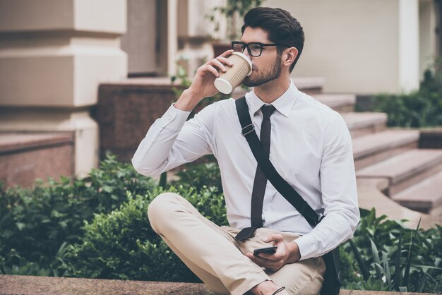 Beba un sorbo de café recién hecho antes de la jornada laboral. Hombre joven confidente en vasos tomando café y sosteniendo su teléfono inteligente mientras está sentado al aire libre