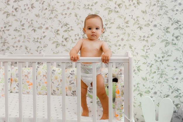 Un bebé de 11 meses en pañal se para en su cuna. niño en la cama.