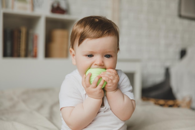 El bebé de 6 meses roe la pelota sosteniéndola con las manos sentado en la cama dando dientes