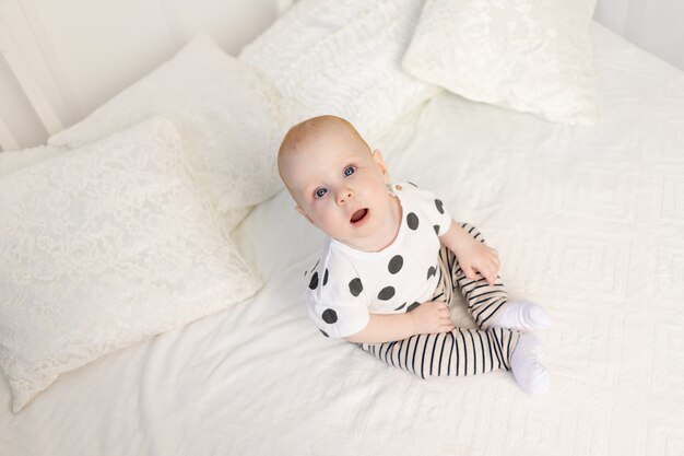 Bebê 8 meses sentado na cama em casa de pijama e olhando para a câmera, vista superior, lugar para texto