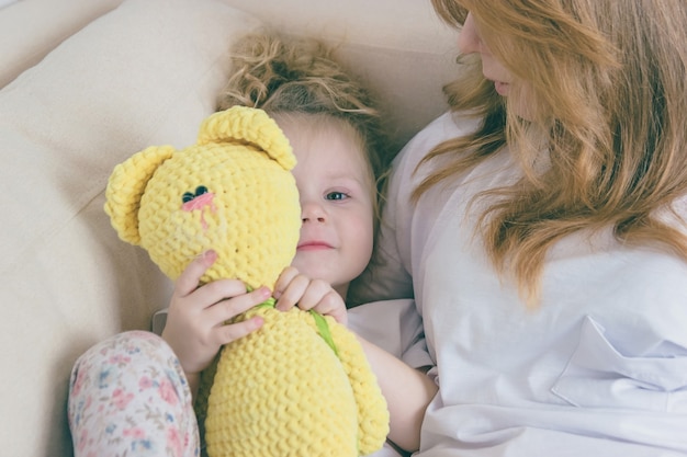 El bebé abraza un peluche. Mamá e hija están en el sofá.