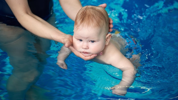 Bebé activo nadando por primera vez en las manos de la mujer en la piscina azul Manos femeninas recortadas sosteniendo cuidadosamente la cabeza del bebé