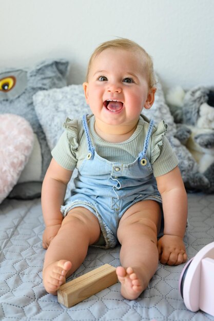 Bebê adorável em roupas casuais sorrindo enquanto está sentado na cama macia com bloco de brinquedo olhando para a câmera em casa