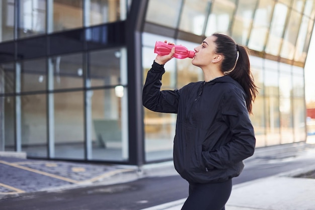 Bebe agua y se toma un descanso La joven morena con ropa deportiva tiene un día de fitness al aire libre en la ciudad