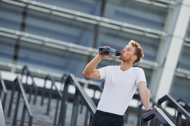 Bebe agua y toma un descanso El joven con ropa deportiva hace ejercicio al aire libre durante el día