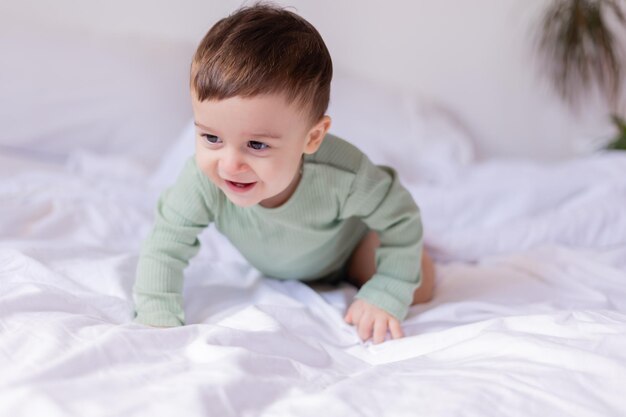 Un bebé alegre con un traje de algodón verde está acostado boca abajo en una cama blanca