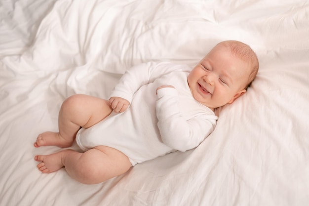 Un bebé alegre con un traje blanco se ríe acostado boca arriba sobre ropa de cama blanca estilo de vida niño feliz