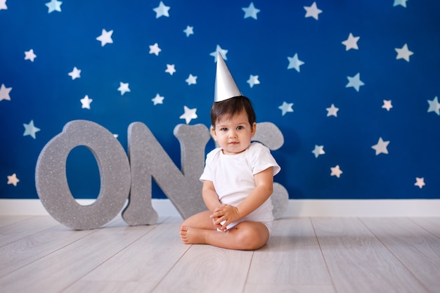 El bebé de un año celebra su cumpleaños cerca de letras plateadas UNO sobre fondo azul con estrellas.