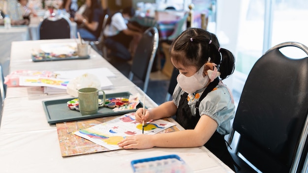 Bebê asiático bonitinho usando máscara sorrindo pintando com tintas coloridas usando aquarela Menina asiática usando pincel desenhando cor Conceito de estilo de vida de atividade do bebê Conceito de distância social