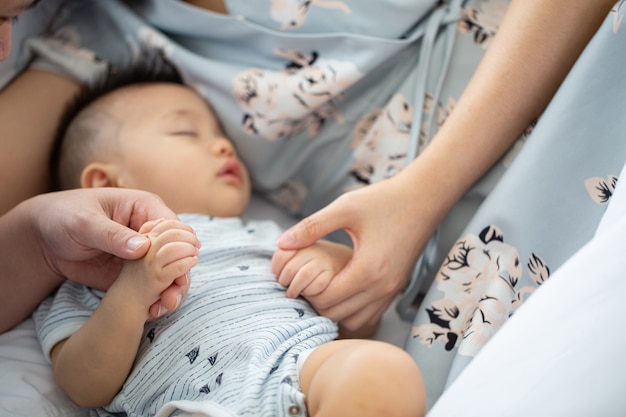 Bebê asiático dormindo com prerent, de quem está segurando suas mãos na cama.