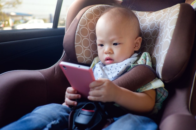 Bebé asiático del niño del bebé que se sienta en el asiento de carro y que mira un vídeo del teléfono elegante