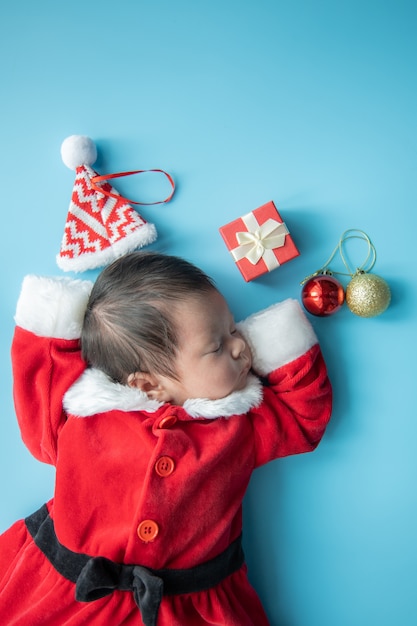 Bebé asiático recién nacido en uniforme de Papá Noel durmiendo con caja roja presente y sombrero rojo sobre fondo azul.