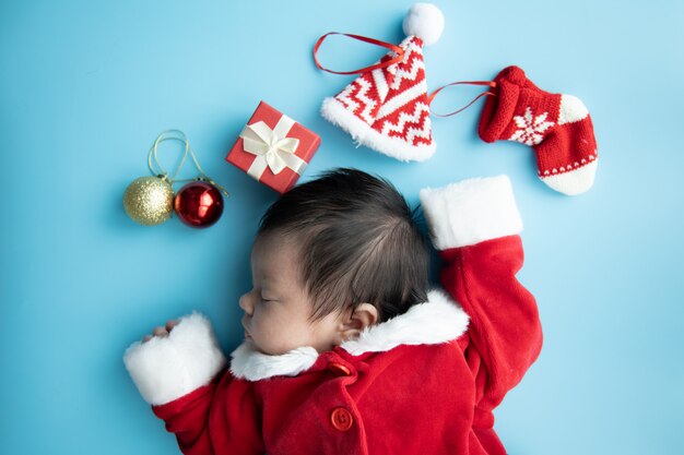 Bebé asiático recién nacido en uniforme de Papá Noel durmiendo con caja roja presente y sombrero rojo sobre fondo azul.