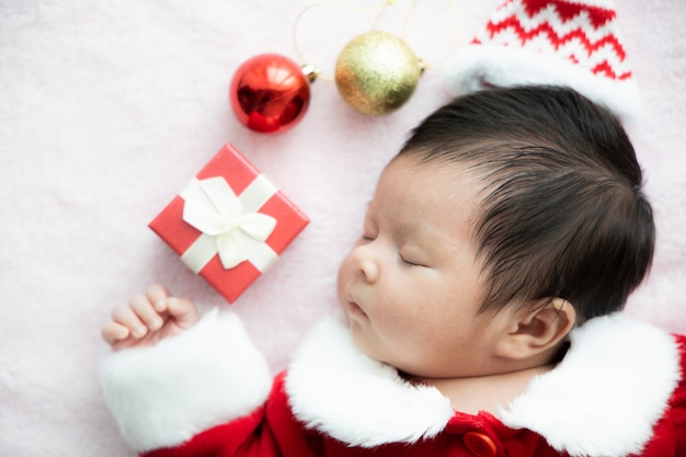 Bebé asiático recién nacido en uniforme de Papá Noel durmiendo con caja roja presente y sombrero rojo