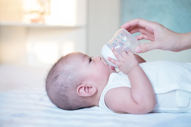 Bebê asiático vestido de branco está deitado na cama bebendo leite de uma garrafa com a mão da mãe