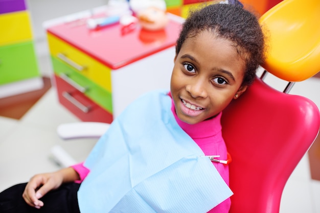 Bebé bonito preto sorrindo sentado em uma cadeira odontológica vermelha