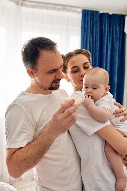 El bebé en los brazos de los padres bebe comida para bebés del biberón, mamá y papá cuidan de su adorable tarifa infantil