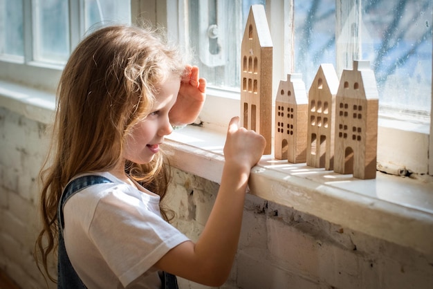 Bebê brincando com casa de madeira Brinquedos ecológicos para crianças, garota branca perto da janela está construindo