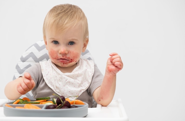 Foto bebé caucásico de aproximadamente 1 año de edad comiendo verduras frescas en silla alta sobre fondo blanco autoalimentación para niños idea de destete babyled nutrición saludable de alimentos sólidos para bebés espacio de copia para texto