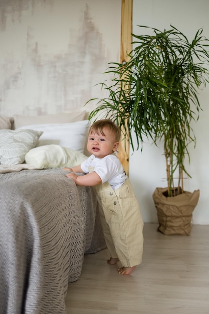 Un bebé caucásico con overoles beige está parado en el suelo cerca de la cama en la habitación El niño aprende a caminar alrededor de la cama