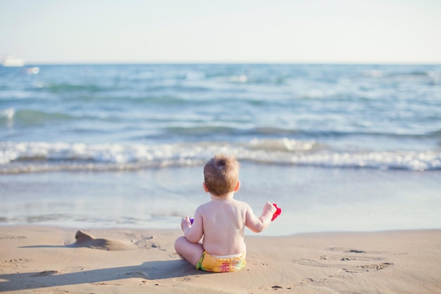 Bebé caucásico sentado en la playa cerca del mar y jugando con juguetes para la arena