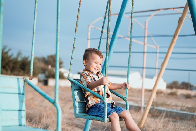 Bebé en columpio balanceándose cerca del mar, en camisa y pantalones cortos de mezclilla