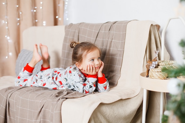 Bebê com cabelo loiro no básico de pijama de papai noel no sofá e sonhando acordado ano novo natal
