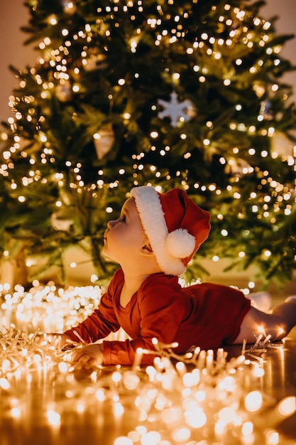 Bebê com chapéu de Papai Noel, deitado de barriga no chão, em uma árvore de Natal com luzes de fundo, olhando para cima.